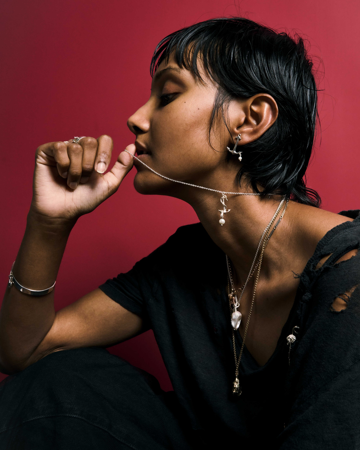A model in profile against a red backdrop, wearing a pearl drop earring, a pearl ear cuff and layered silver chains with a baroque pearl pendant