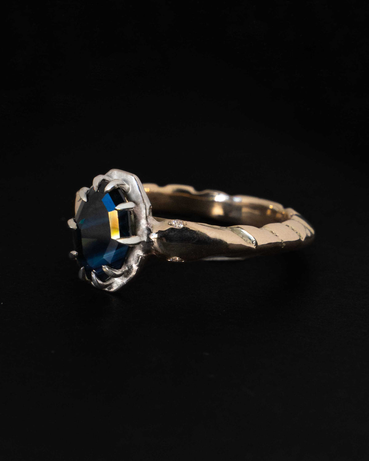 A teal parti sapphire bezel-set in solid gold between two kite-cut diamonds, on white satin