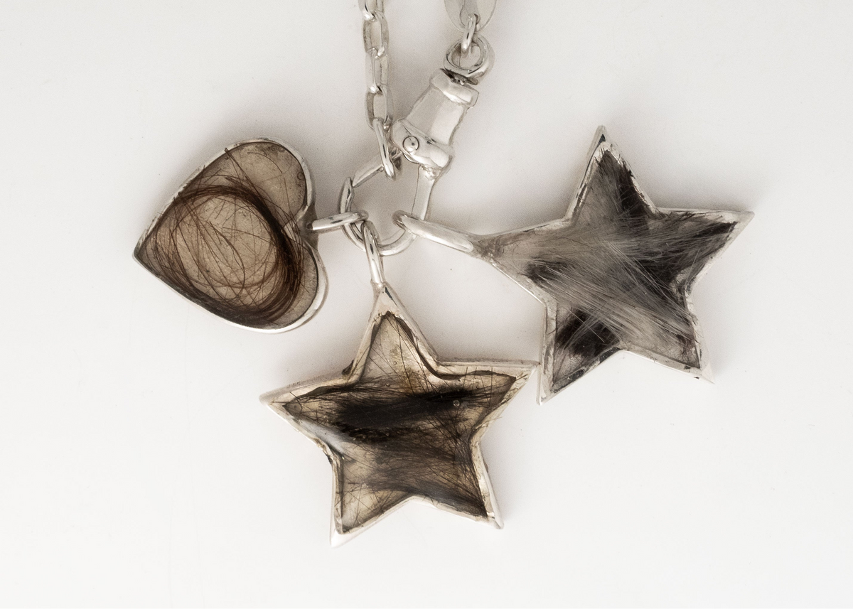 Three sterling silver heart pendants on black: two hold a lock of hair under a clear setting, the third is engraved MUM