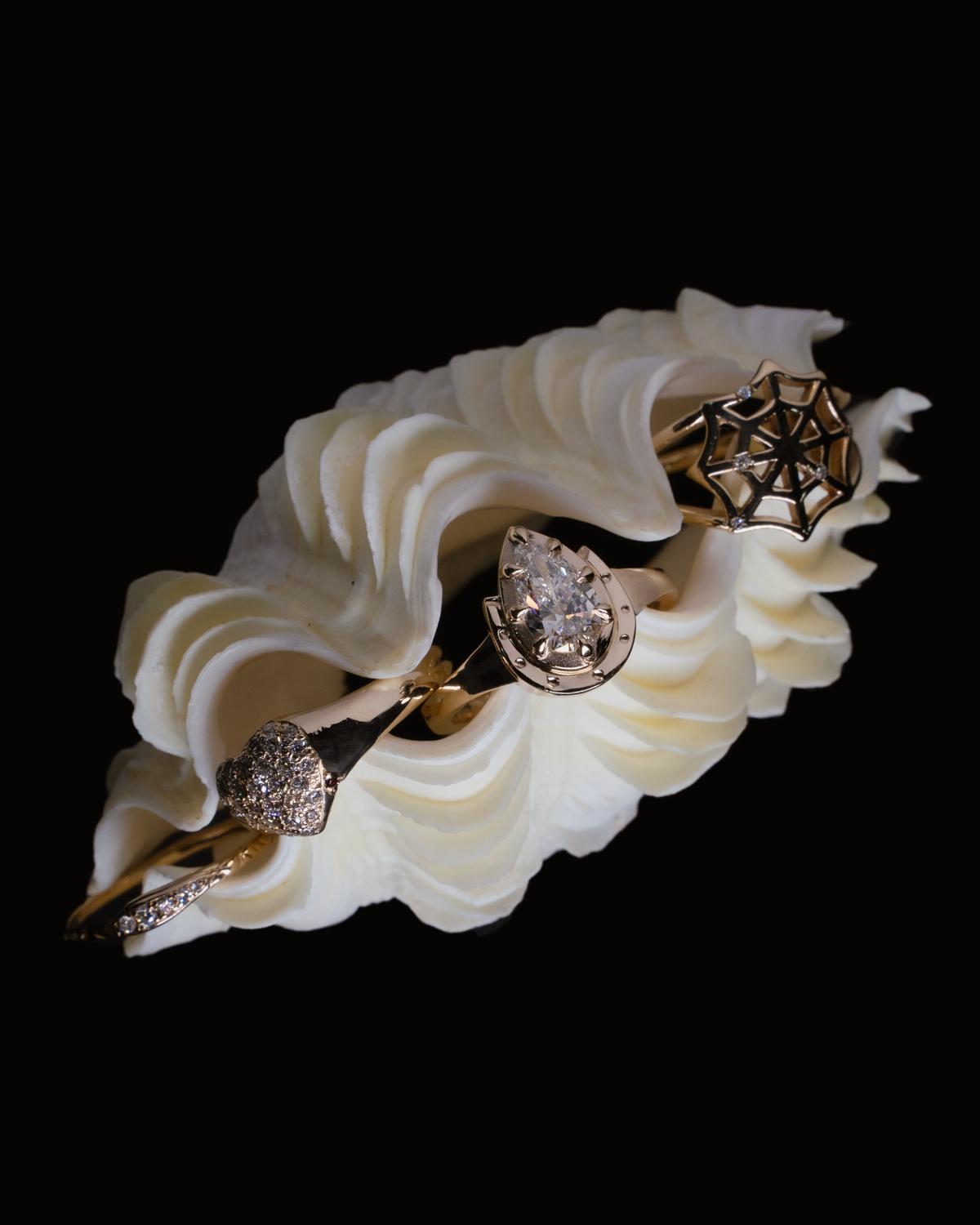Three solid gold rings resting in a fluted white shell: a pave diamond heart, a pear-cut diamond in a horseshoe setting and a diamond-set spider web, on black