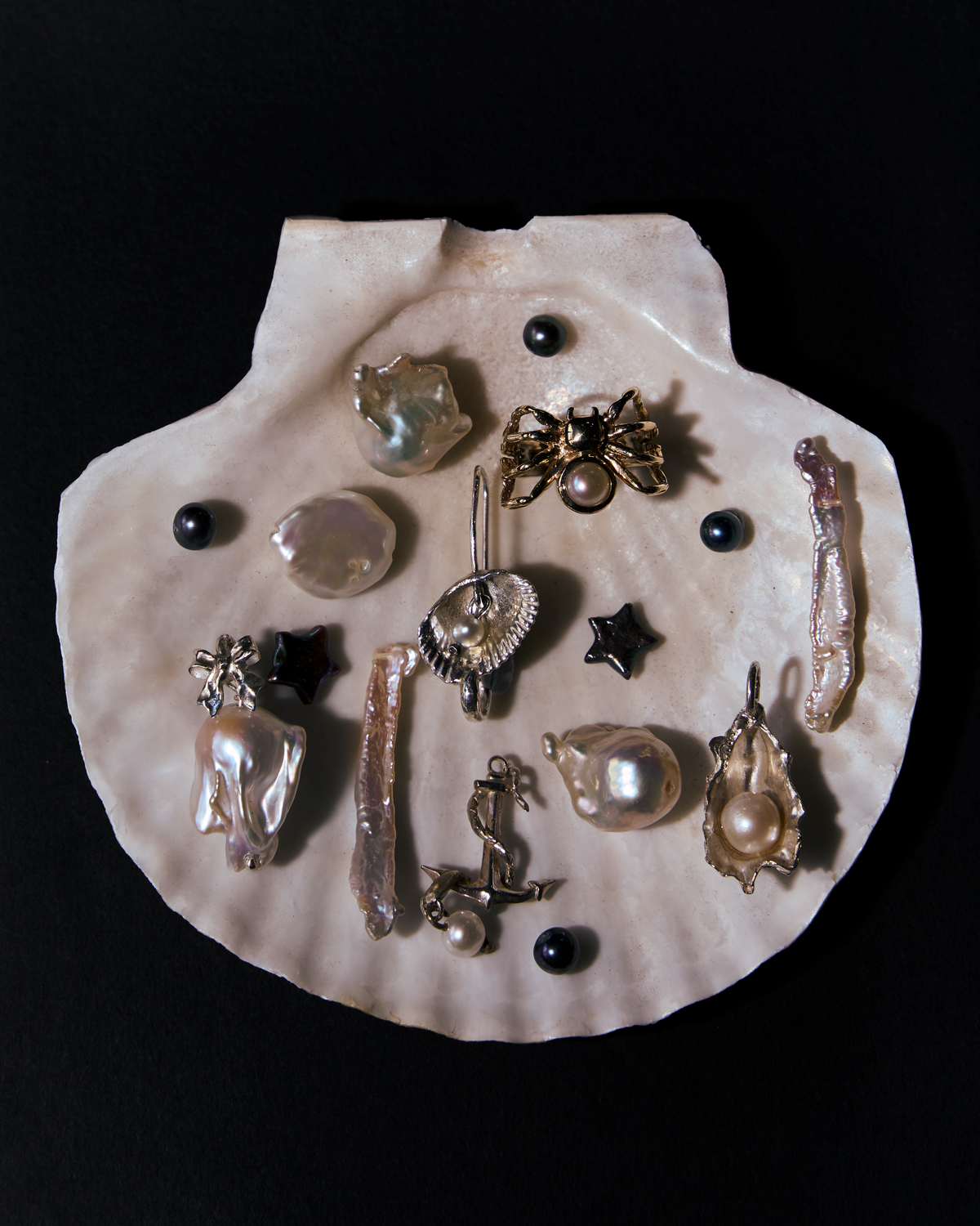 A scallop shell holding loose baroque and keshi pearls, black pearls and star charms, with silver and gold pearl-set pendants: a spider, an anchor, a shell and an oyster, on black