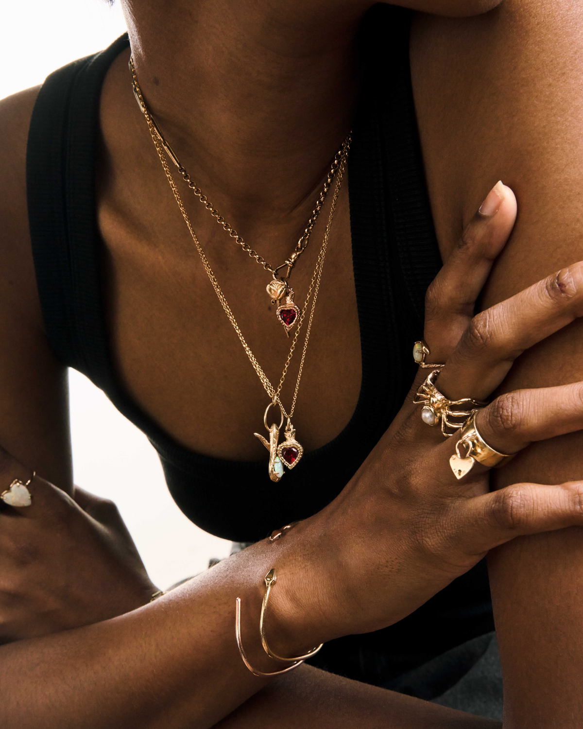 A model in a black singlet wearing layered gold chains with a garnet heart, a serpent and opal charm, gold rings set with white opals, a spider ring and a heart padlock ring, and gold bangles