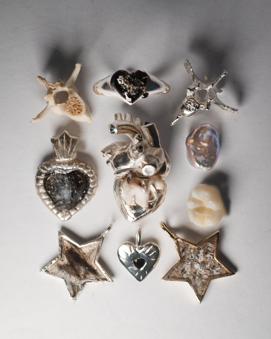 Sterling silver memorial pieces laid out on white: a signet ring and a sacred-heart pendant set with cremation ashes, two star pendants holding ashes, an anatomical heart, a small heart charm, a tooth, a pearl and two vertebrae