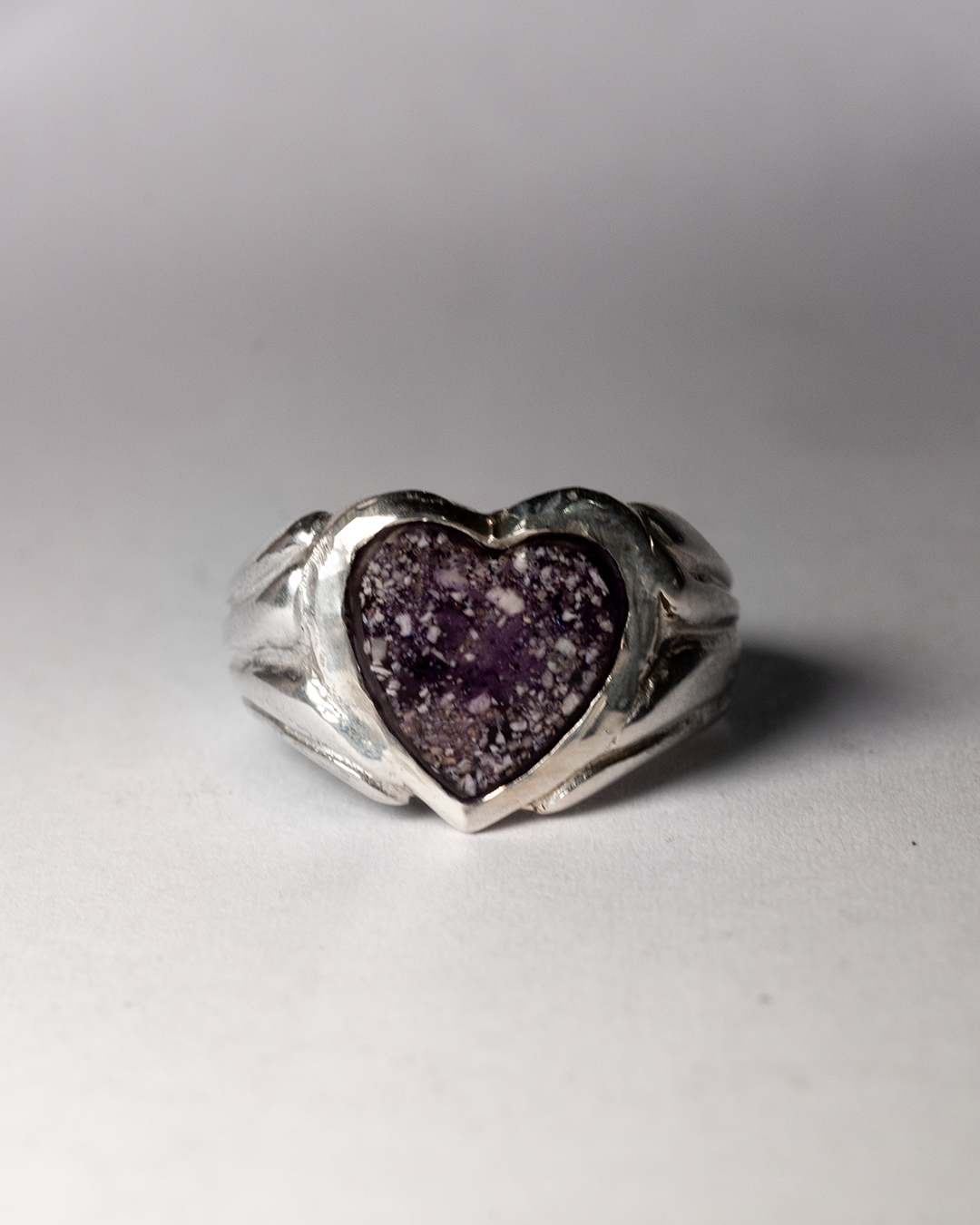 A sterling silver heart ring with ashes sealed over purple enamel, on a white background